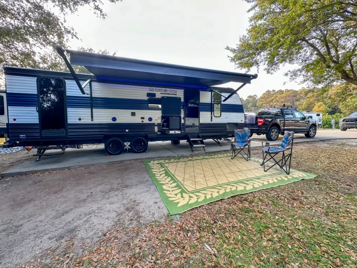 Sunny RV rental setup: camper with extended awning, blue lights, outdoor mat, chairs, & pickup at campsite.. Forest River Grey Wolf 26 DBH 2025