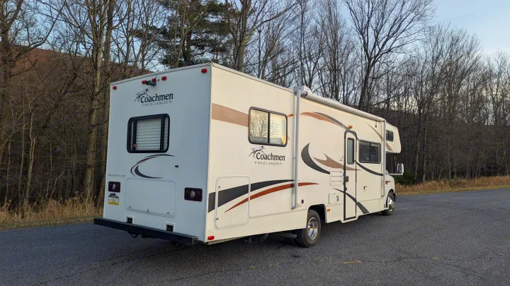 White Coachmen Freelander RV on asphalt, backed by bare trees & a hill. Perfect for an outdoor adventure or rv rental.. Forest River Coachmen Freelander 2013