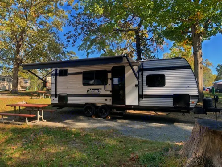 This RV rental, a Coleman travel trailer, is set up at a sunny campsite with a picnic table & awning extended.. Keystone Coleman 23B 2025