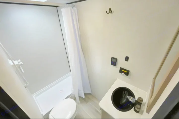 An RV rental's compact bathroom: featuring a white shower with curtain, toilet, and dark sink vanity on a light floor.. Keystone Coleman 23B 2025