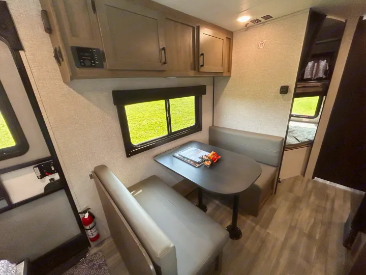 An RV rental features a compact dinette with grey benches, a dark table, overhead cabinets, and a window view. Bunks are visible.. Starcraft Autumn Ridge 2025