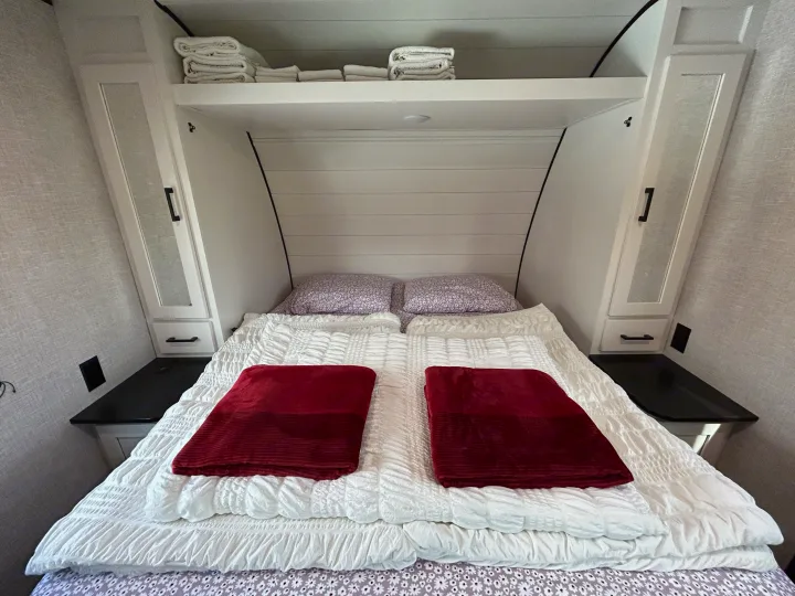 Cozy RV rental bedroom with white bedding, red throws, and built-in storage cabinets above and beside the bed.. Starcraft Autumn Ridge 2025