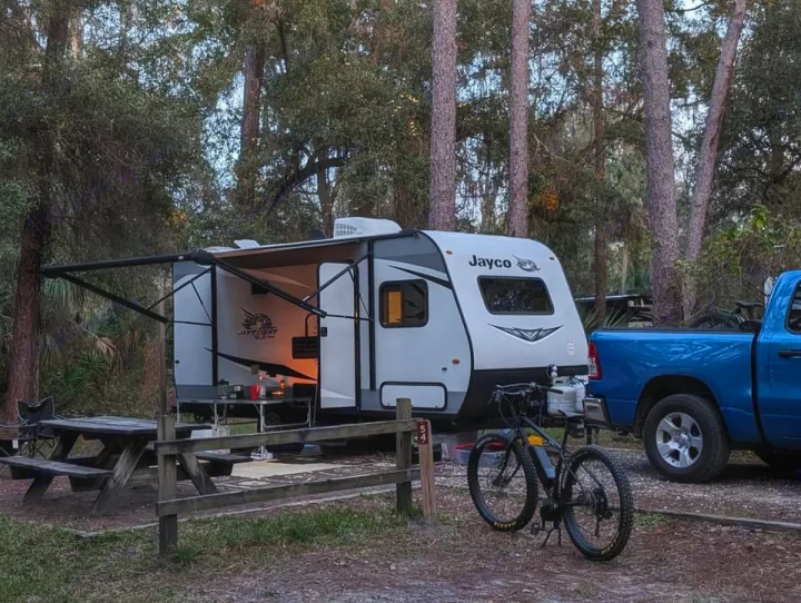 Campsite setup: Jayco RV rental with awning extended, blue truck, and mountain bike in a wooded outdoor setting.. Jayco Jay Flight SLX 2021