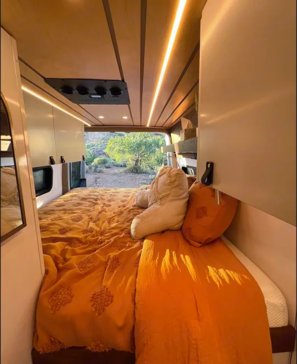 Step into this cozy RV rental's bedroom, featuring a vibrant orange bed, warm ceiling lights, and stunning outdoor views.. Mercedes Sprinter Van 170 4x4 2022