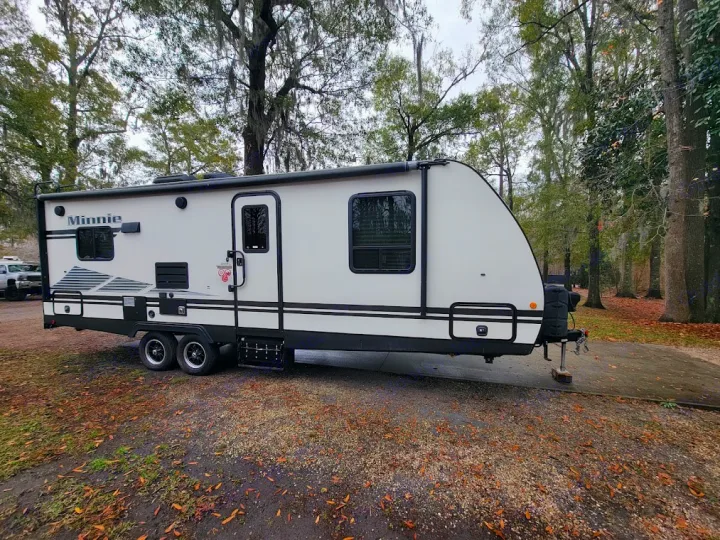 A white Minnie travel trailer RV rental is parked on a gravel site, surrounded by trees. Ready for your camping trip!. Winnebego Minnie Winnie 2020