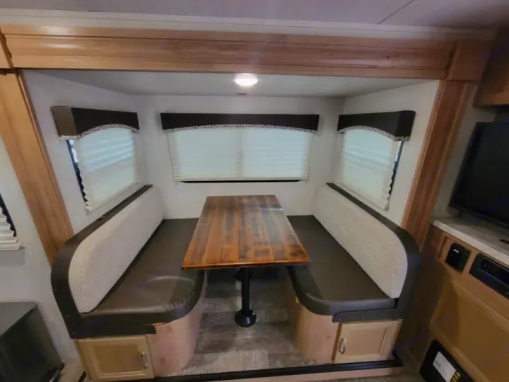 Interior of an RV rental: A cozy dinette with two facing cushioned benches, a wooden table, and windows with white blinds.. Winnebego Minnie Winnie 2020