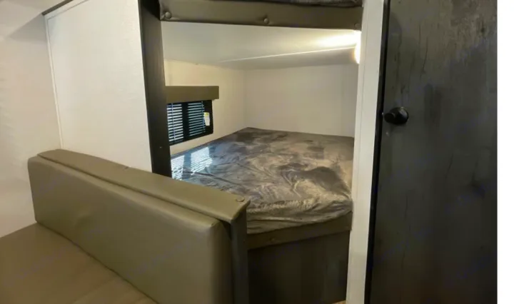 RV rental interior: View of a compact bedroom with a large bed, upper bunk, window, adjacent sofa, and a dark door.. Keystone RV Hideout 2023