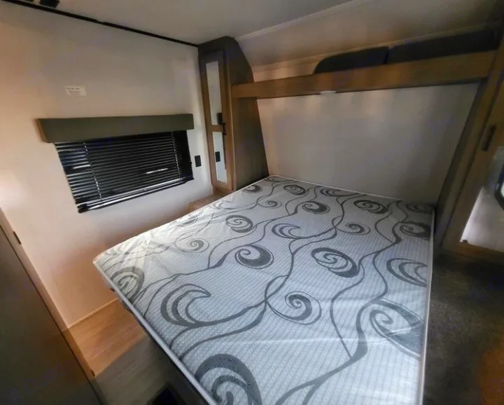 RV rental bedroom with a comfortable bed, window with blinds, and built-in storage in a travel trailer.. Keystone Hideout 2021