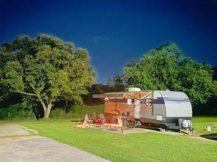 An RV rental shines at a night campsite. Its awning is extended over a warm fire pit and chairs under the starry sky.. Avenger LE 2022