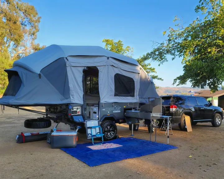 A gray pop-up rv rental camper and black SUV parked at a dirt campsite with an outdoor kitchen setup.. Air Opus OP4 2021