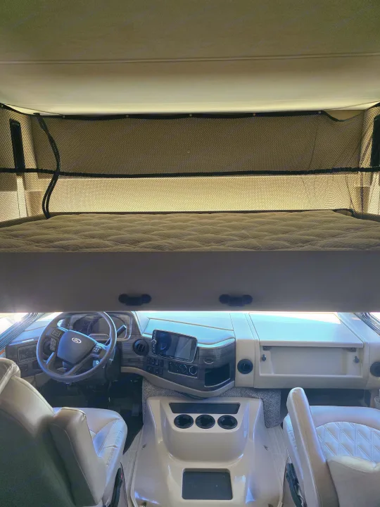 Interior of an RV rental: driver's cockpit with dashboard, steering wheel, and an overhead bunk bed with safety net.. Thor Challenger 2021