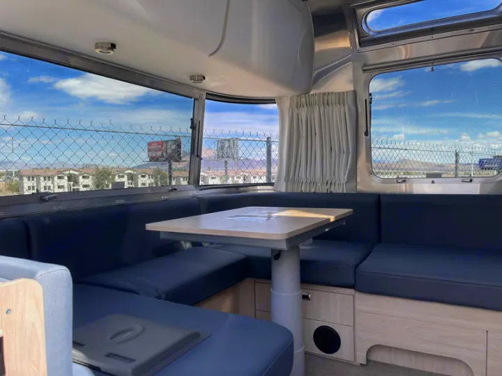 Spacious RV rental interior: Airstream-style dinette with blue seating & table. Wide windows reveal a city and cloudy sky view.. Airstream Globetrotter 2024