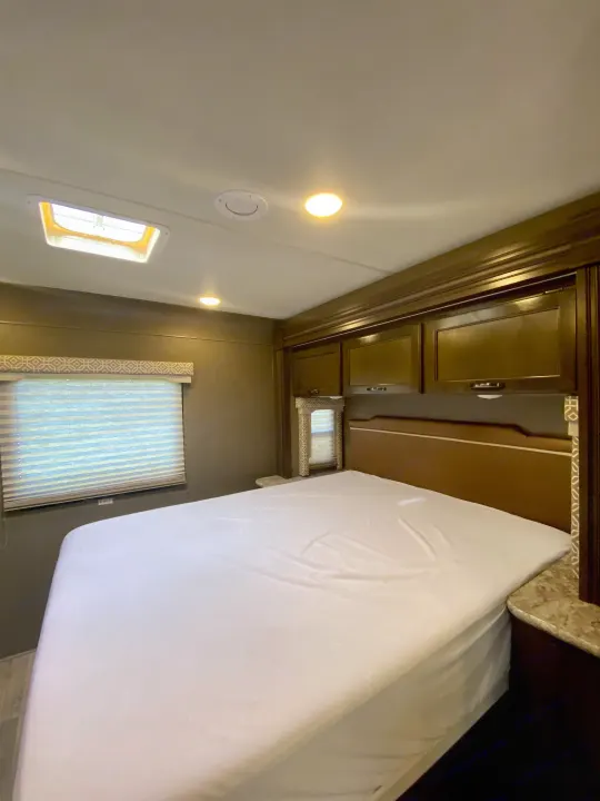 RV rental bedroom: a clean, white-sheeted bed, window with blinds, and dark wood overhead cabinets.. Thor Freedom Elite 2018