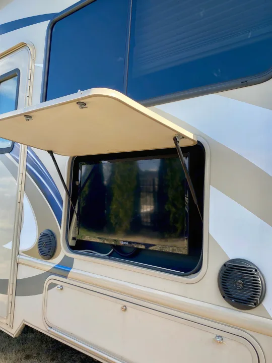 RV rental with outdoor TV. Side view of a beige RV with an open compartment revealing a large screen TV and speakers.. Thor Freedom Elite 2018