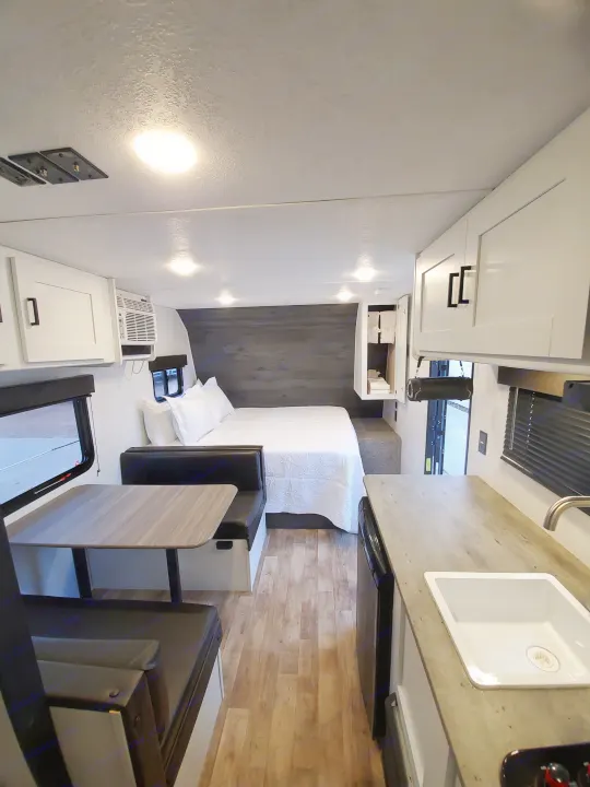 Clean and modern RV rental interior showing a bedroom with a white bed, dinette area, and compact kitchenette.. Keystone RV Hideout 2021