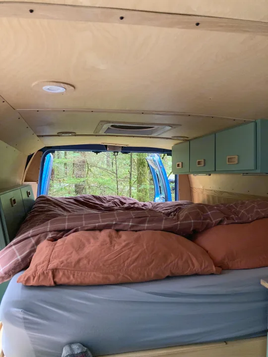 Inside an RV rental: a cozy bed with brown & orange bedding, overhead cabinets, and a scenic forest view.. Ford Econoline 2006