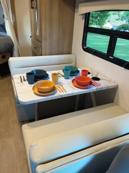 Cozy RV rental dinette with a marble table set with colorful dishes, surrounded by comfy bench seating, and a glimpse of the bedroom.. Nexus Phantom 2025