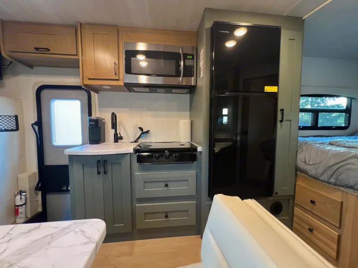 RV rental interior: A compact kitchen with black refrigerator, stainless microwave, stovetop, and sink. Light wood and gray cabinets, plus a bed in background.. Nexus Phantom 2025