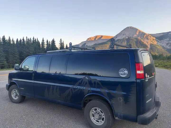 A dark blue RV rental van with mountain decals is parked by a road, backed by evergreen trees and majestic mountains.. Chevrolet Express 2003
