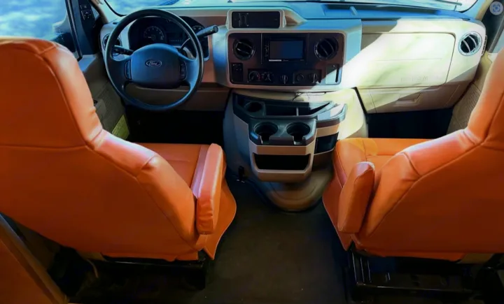 Inside an RV rental: wide view of the front cabin with two orange seats, Ford steering wheel, and a functional dashboard.. Winnebago Minnie Winnie Premier 2015