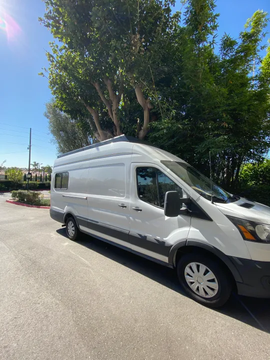 White Ford Transit RV rental van parked on sunny asphalt beside green trees.. Ford Transit 2021