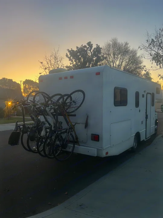 White RV rental with bikes on a rear rack, parked on a street. Early morning light casts silhouettes against the orange-blue sky.. Thor Majestic 2019