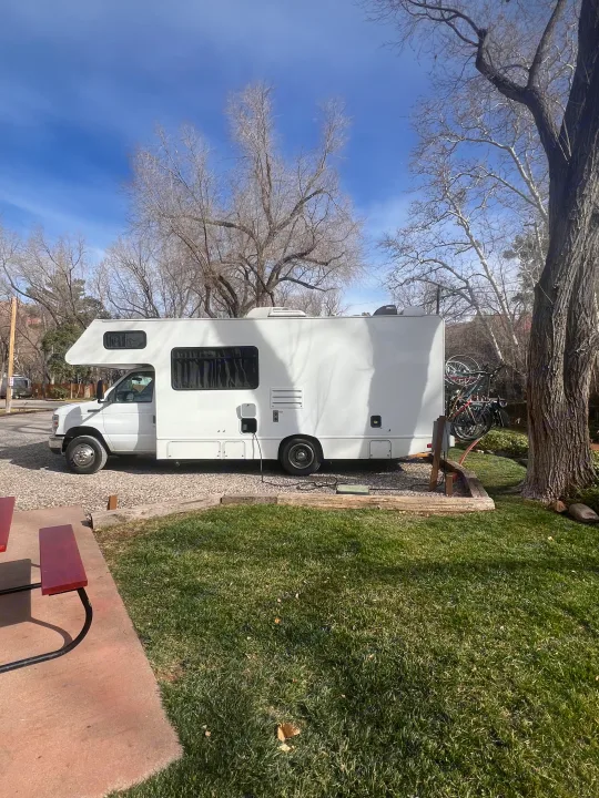 White RV rental at a sunny campsite, equipped with bikes. Green grass, bare trees, and blue sky in the background.. Thor Majestic 2019
