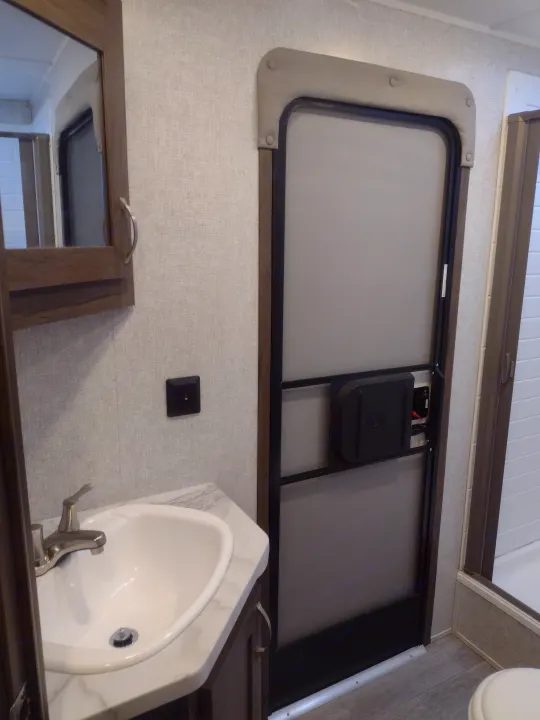 Modern RV rental bathroom featuring a vanity with sink, mirror, and compact shower stall, alongside a main interior door.. East to West Silver Lake 2021