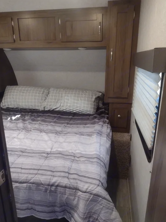 Cozy rv rental bedroom features a comfy bed with striped comforter & checkered pillows, storage cabinets, & window blinds.. East to West Silver Lake 2021