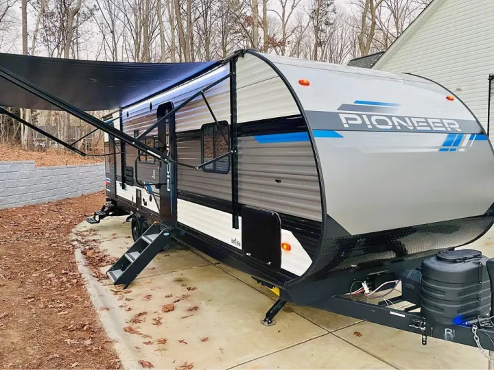 A grey and black Pioneer travel trailer RV with a black awning extended, parked on a paved driveway. Ideal for an rv rental.. Heartland RVs Pioneer 2023