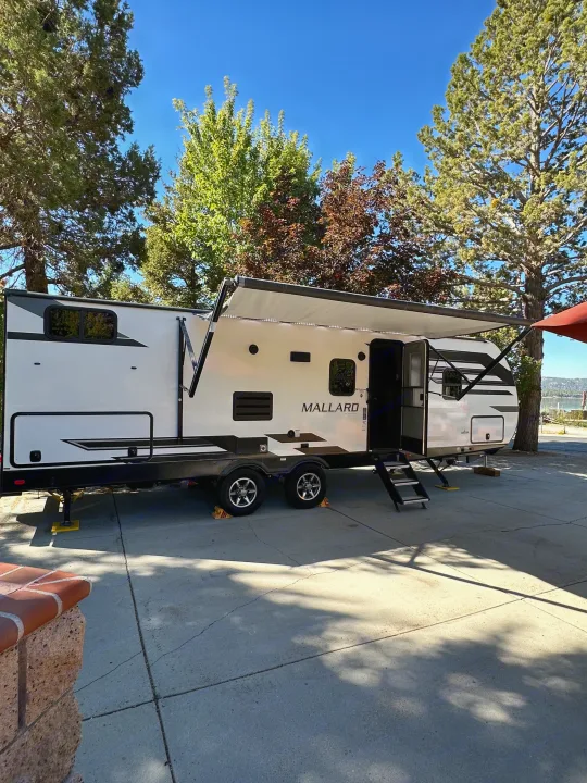 White RV rental with awning extended and steps down, parked on a paved site by a lake for vacation.. Heartland RVs Mallard 2022