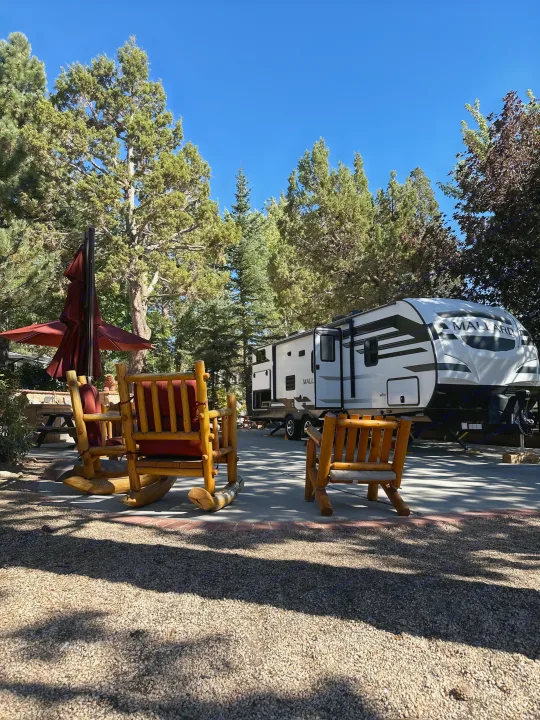 An RV rental travel trailer parked at a sunny campsite, surrounded by tall trees and inviting wooden chairs.. Heartland RVs Mallard 2022