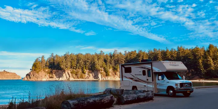 A beige Winnebago Minnie Winnie RV rental parked by a tranquil bay. Dense forests line the distant shore under a bright blue sky.. Winnebago Minnie Winnie 2017