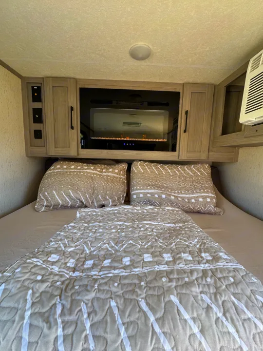 A modern RV rental bedroom featuring a bed with patterned bedding, overhead cabinets, an electric fireplace, and an AC unit.. Dutchmen Other 2023