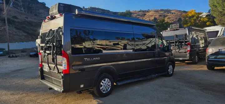 Dark gray Tellaro Ram Promaster RV rental camper van, equipped with a bike rack, in a sunny, dusty lot.. Thor Tellaro 2022