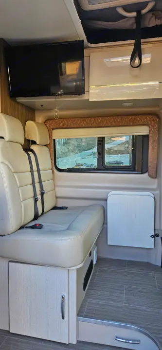 Inside an RV rental: a modern interior with plush bench seating, mounted TV, window views, and a wall-mounted table.. Thor Tellaro 2022
