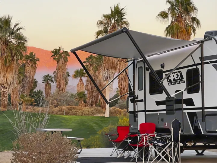 Experience serene desert camping! This RV rental is set up with chairs & awning, bathed in sunset hues, with palm trees.. Coachmen Apex Nano 2023