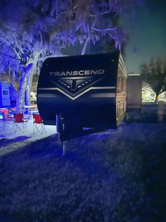 Nighttime view of a Transcend RV rental at a campsite, with blue lights on trees and chairs. Ideal for camping.. Grand Design Transcend Xplor 2024