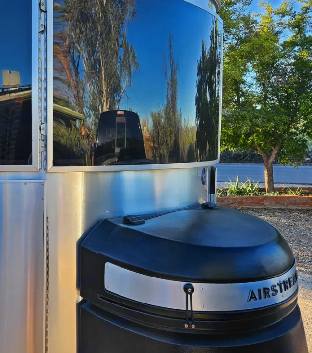 Front of a polished Airstream RV rental, reflecting blue sky and trees. Its silver body contrasts with a black tank cover.. Airstream Basecamp 2020