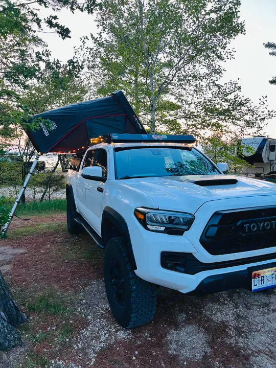 White Toyota Tacoma truck with an iKamper roof-top tent, set up for camping next to an RV rental by the water, for an overlanding adventure.. Toyota Tacoma TRD-Pro 2020