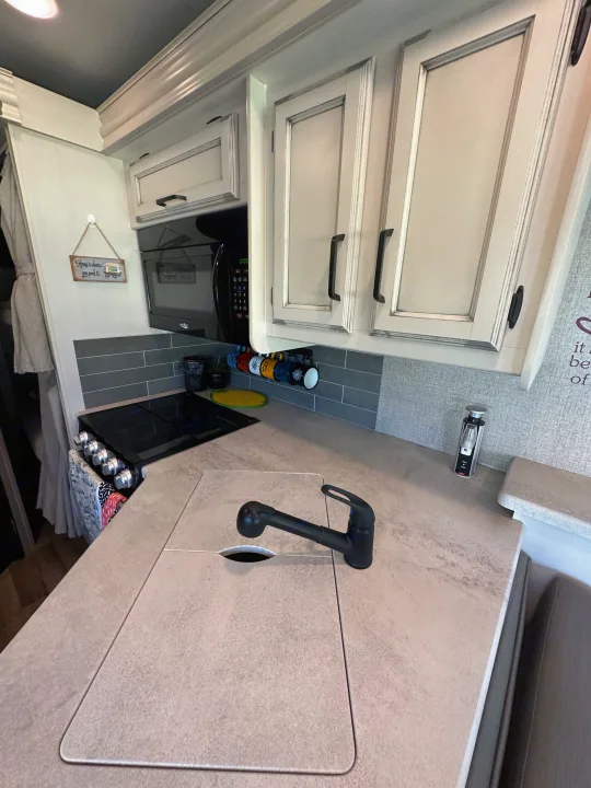 A modern RV rental kitchen featuring light cabinets, a black stove, microwave, and a compact counter with a covered sink.. Entegra Coach 29F 2021