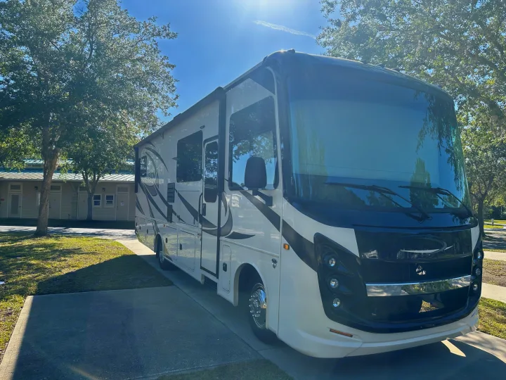 Front-view of a modern white and black rv rental, parked on asphalt with trees & building in bright sunlight.. Entegra Coach 29F 2021