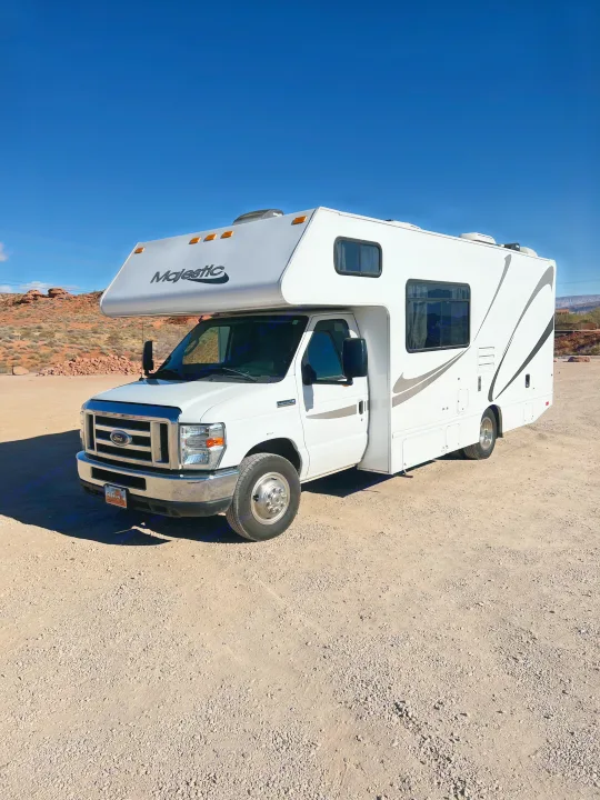 A white Majestic RV rental is parked on a dusty road, set against a backdrop of red rock hills under a clear blue sky.. Thor Majestic 2018