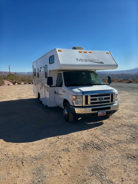 A white Majestic RV rental is parked on a sunny dirt lot with mountains and a town in the distance under a blue sky.. Thor Majestic 2018