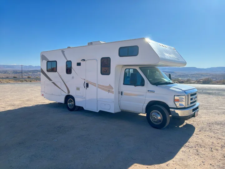 White Majestic motorhome RV rental on an unpaved desert lot with mountains & town under a clear blue sky.. Thor Majestic 2018