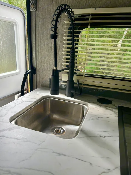 Inside an RV rental: a modern kitchen with a black gooseneck faucet, stainless sink, and marble-look counter.. Nexus RV Phantom 2023