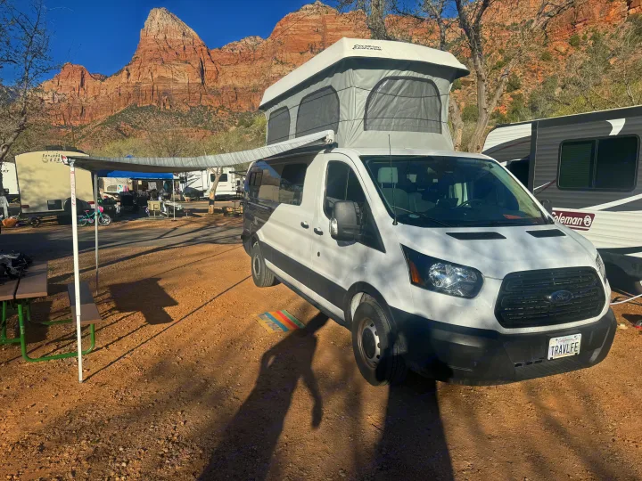 White camper van (an RV rental) with a pop-top, parked at a sunny campground against stunning red rock mountains.. Ford Transit Custom 2019
