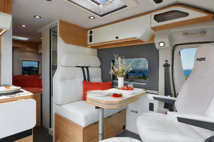 Inside a modern RV rental, featuring a comfortable dining area with a coastal view, kitchen, and bedroom beyond.. Pilote V600G EVIDENCE 2025