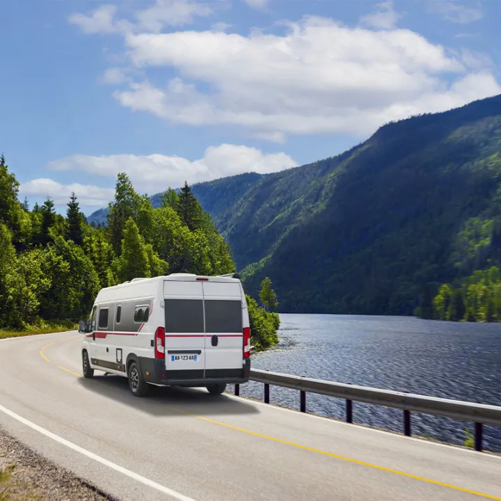 This image shows a white RV driving on a winding road beside a calm lake with mountains and forests. Perfect for an RV rental adventure!. Pilote V600G EVIDENCE 2025