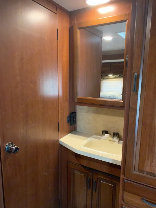 RV rental bathroom with sink, countertop, mirror, and wood cabinets. Door to left, bedroom reflected in mirror.. Forest River Sunseeker 2014